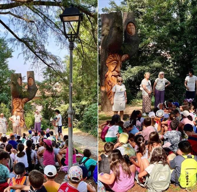 Bambini seduti ad ascoltare vicino alle sculture di legno del parco Belvedere